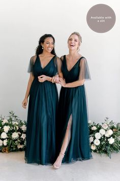 two women standing next to each other in long dark green dresses with sheer sleeves and thigh high slits