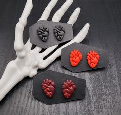 three pairs of heart shaped earrings sitting on top of a table next to a skeleton's hand