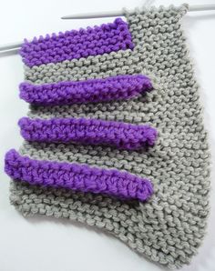 three crocheted purple and gray pieces of cloth on a white table with knitting needles