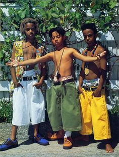 three young boys standing next to each other in front of a white fence and green plants