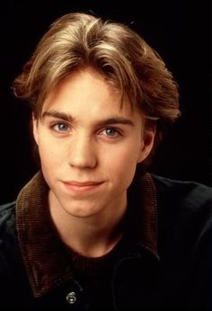 a young man with blue eyes looks into the camera while wearing a black shirt and brown jacket