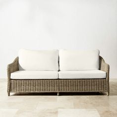 a wicker couch with white pillows on it in front of a wall and tiled floor