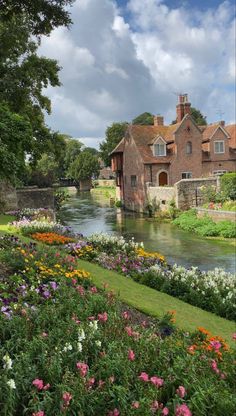 a river running through a lush green field next to a building and flowers growing on the side of it