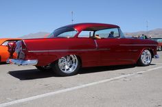 an old red car parked in a parking lot next to other classic cars and people