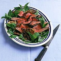 a white plate topped with meat and greens next to a knife on top of a blue table cloth