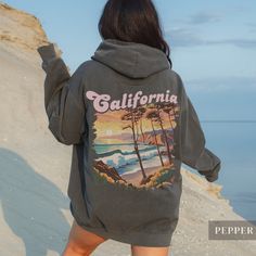 "Our Vintage California Sunset Hoodie - Embrace the essence of California with our cozy and luxurious hoodie featuring a striking retro design on the back.  With \"California\" in bold, distressed text overlaid on a mesmerizing art deco-style beach scene, complete with crashing waves, windblown trees, and a radiant sunset, this hoodie exudes West Coast vibes!  Crafted for ultimate comfort and warmth, it feels like an old favorite from the very first wear, making it a must-have for those who adore California's laid-back atmosphere. Whether you're a local or simply enamored with the Golden State, this hoodie serves as a cherished memento or a thoughtful gift for any occasion. .: 80% Ringspun US cotton, 20% Polyester .: Heavy fabric (9.5 oz /yd²) .: Relaxed fit - Please reference the size cha Oversized Long Sleeve Vsco Hoodie, Oversized Vsco Style Hoodie, Oversized Vsco Sweatshirt For Streetwear, Vsco Style Winter Hoodie For Streetwear, Vsco Winter Hoodie For Streetwear, Oversized Vsco Sweatshirt For Winter, Oversized Graphic Print Hoodie For Outdoor, Oversized Hoodie With Graphic Print For Outdoor, Long Sleeve Vsco Sweatshirt For Loungewear