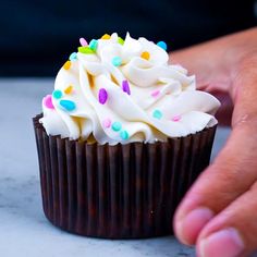 a cupcake with white frosting and sprinkles being held by someone's hand