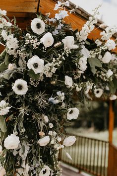 an arrangement of white flowers and greenery hanging from a wooden structure