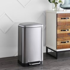 a stainless steel trash can next to a wooden chest with flowers on it and a vase in the corner