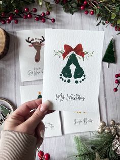 someone holding up some christmas cards on top of a table with other holiday decorations and greenery