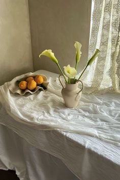 a white vase with flowers and oranges on a table next to a curtained window