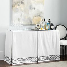 a white table with bottles and glasses on it next to a chair in front of a painting