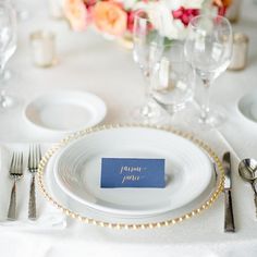 a place setting with white and gold plates, silverware, and flowers in the background