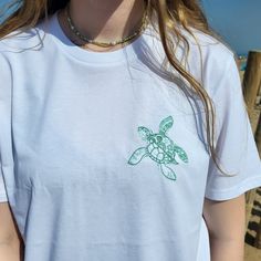 This is a personalised, white embroidered t-shirt with our Turtle image on the front.  Please select the personalisation tab below to choose the thread colour of your choice and to add any matching colour text to the arm up to a max of 12 characters, such as initials, a memorable beach, a name or a location.   A great addition to your wardrobe or as a gift the high quality great fitting organic unisex t-shirt is perfect for any occasion, not only is it organic but it is also PETA Vegan approved. It's a must-have for anyone who loves the sun, sand, and surf! This ultra-comfy and stylish t-shirt is the perfect gift for beach enthusiasts, ocean lovers, or anyone who appreciates a one-of-a-kind piece of clothing that brings back memories of sun-soaked days, sea or lovely summer holidays. Make Custom Embroidered Summer T-shirt, Summer Beach T-shirt With Custom Print, Embroidered Short Sleeve Beach T-shirt, Turtle T Shirt, Sea Turtle Shirt, Turtle Images, Turtle Gifts, Embroidered Tshirt, Personalized T Shirts