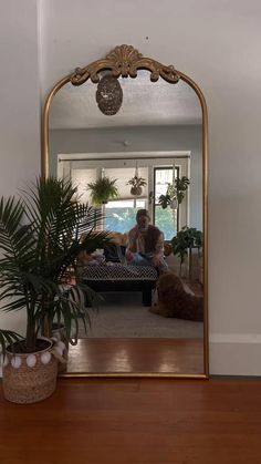 a mirror reflecting a woman sitting on a couch in front of a coffee table and potted plant