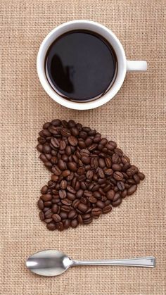 a cup of coffee next to a spoon on a burlap surface with the shape of a heart made out of coffee beans