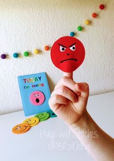 a hand holding up a red paper cutout with an angry face on it, next to a book