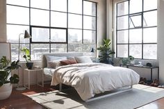 a bedroom with large windows and a bed in the middle, surrounded by potted plants