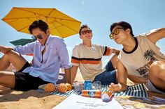 three people sitting on the beach with an umbrella and drinks in front of their faces