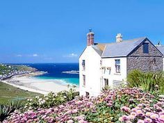 a white house sitting on top of a lush green hillside next to the ocean with purple flowers in front of it