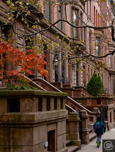 two people walking down the sidewalk in front of some brownstone buildings with autumn leaves on them