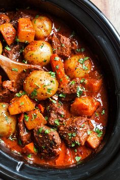 a crock pot filled with meat and potatoes on top of a wooden table next to a spoon