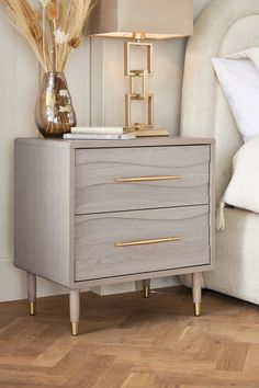 a nightstand with two drawers and a lamp next to it on a wooden floor in front of a white couch