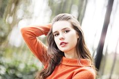 a young woman in an orange sweater poses for a photo with her hand on her head