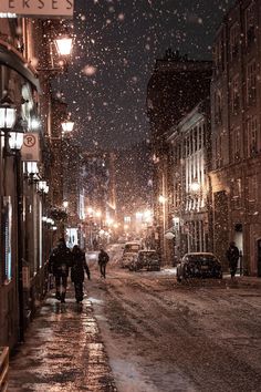 two people walking down a snowy street at night with snow falling on the ground and buildings in the background