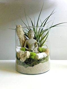 there is a small buddha statue in a glass vase filled with plants and rocks on the table