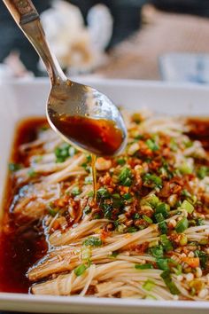 a spoon is pouring sauce over noodles in a white dish with red sauce and green onions