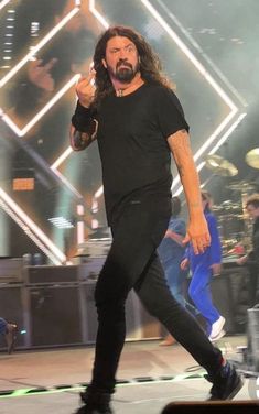 a man with long hair and black pants on stage holding his hand up in the air