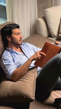 a man sitting on top of a couch holding an open book in his hand and looking at the screen