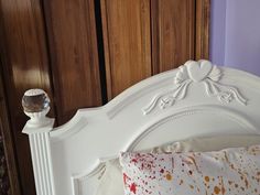 a white bed with an ornate headboard and pillows on it's sides, against a purple wall