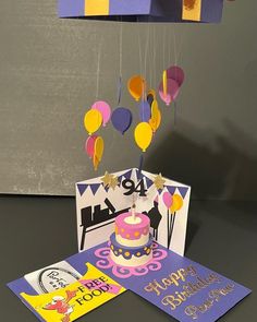 a birthday card with a cake on it and balloons hanging from the ceiling above it