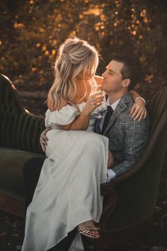 a man and woman sitting on a bench in the woods, one is holding his arm around the other's neck