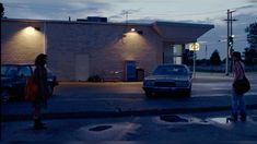 two people crossing the street in front of a gas station at night with cars parked nearby