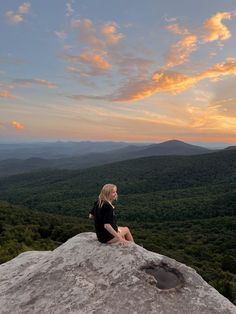 a woman sitting on top of a large rock in the middle of a forest at sunset