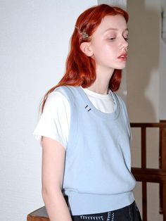 a young woman with red hair wearing a blue shirt and black leather skirt, standing in front of a white wall