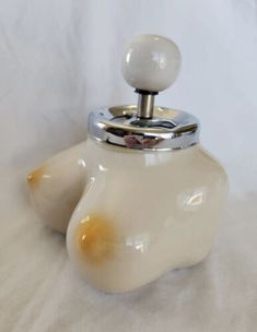 a cream colored tea pot with a metal lid and an egg on the top, sitting on a white surface