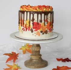 a white cake with chocolate frosting and fall leaves on it sitting on a pedestal