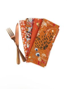 three napkins with different designs on them next to a fork and knife, one has an orange flower design