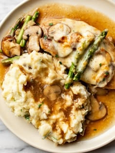 a white plate topped with mashed potatoes covered in mushrooms and asparagus next to gravy