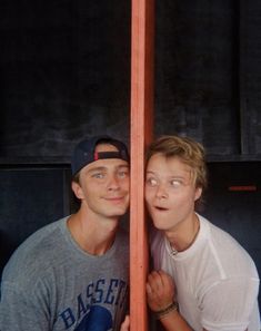 two young men standing next to each other in front of a wooden pole with their eyes closed