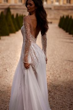 a woman in a long white dress standing on a gravel road with her back to the camera