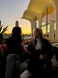 two women sitting on a couch talking to each other in front of a building at sunset