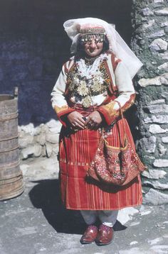 an old photo of a woman dressed in traditional costume and headdress, standing next to a stone wall