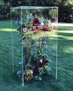 a glass box with flowers and greenery in it on the grass near some trees