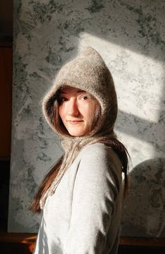 a woman wearing a gray hoodie standing in front of a marble wall with sunlight coming through the window