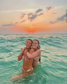 two women in the ocean hugging each other and smiling at the camera with an orange sunset behind them